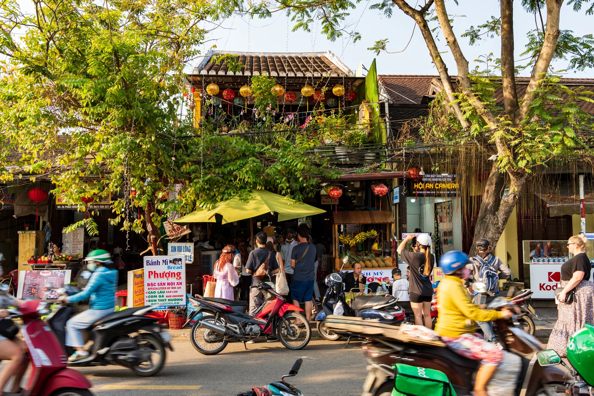 Bánh mì Phượng（ばいんみーふーん）の外観。店が木に覆われており、前の道を忙しくバイクが行き交っている。