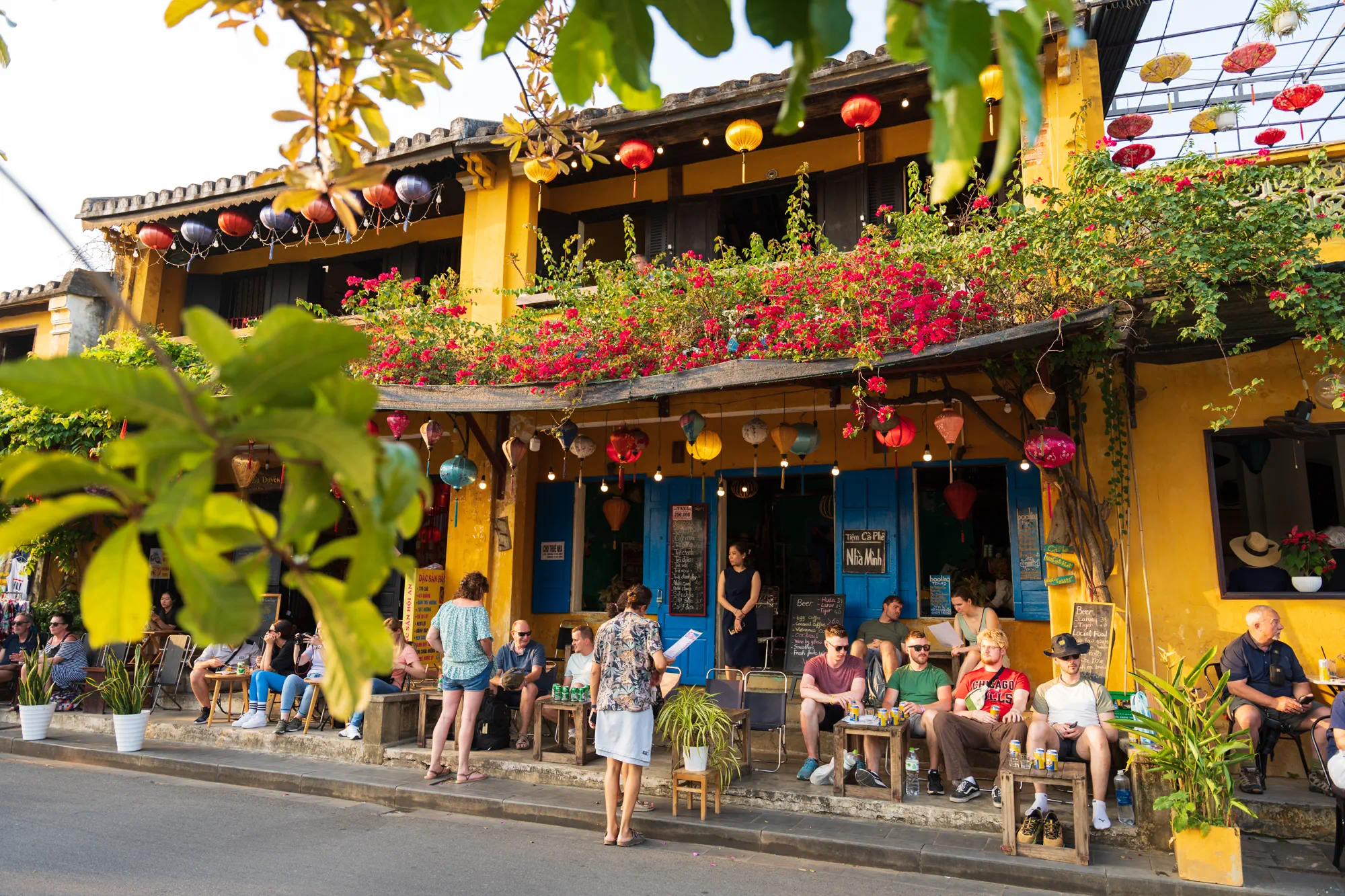 カフェの店頭。木と花に覆われた軒先と、その下で飲食する観光客