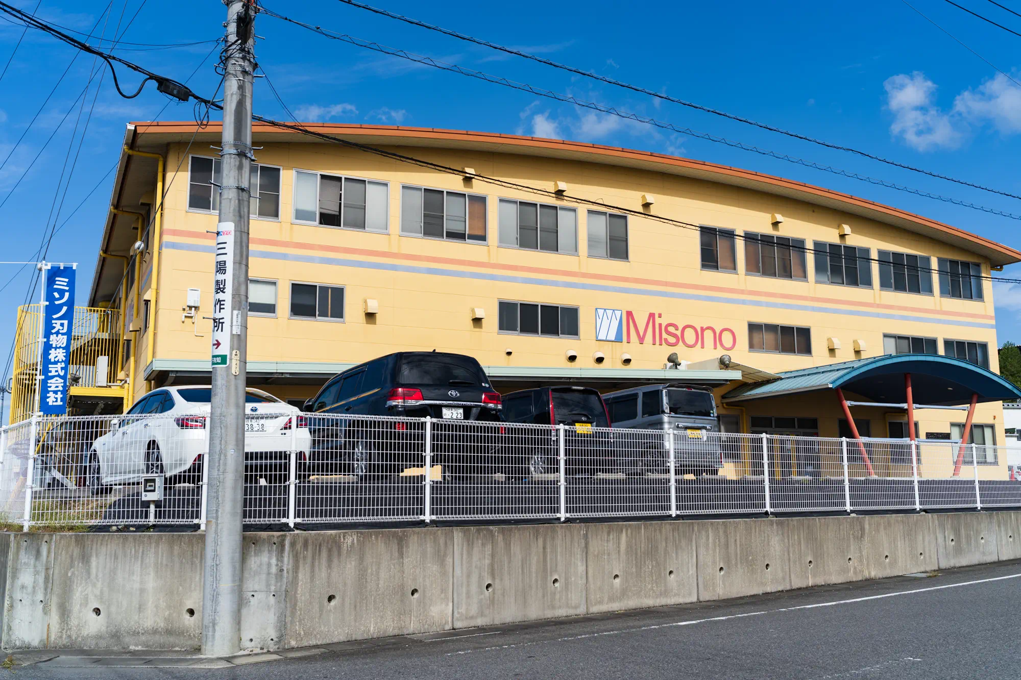 ミソノ刃物の本社工場遠景。黄色い3階建ての社屋と駐車場。手前に青色の社名入りののぼりが立っている